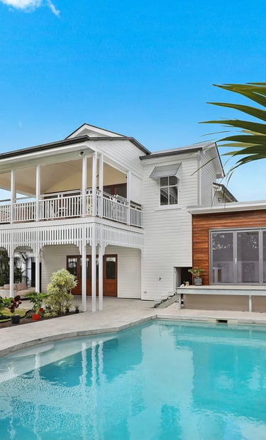 Modern luxury home exterior featuring a white two-story deck, backyard swimming pool, and tropical landscaping.