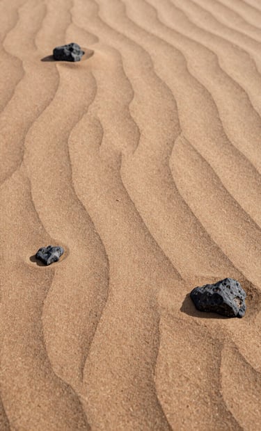 Detailed photographic shot of natural sand ripples and small desert rocks. The lighting is soft and warm, highlighting the natural beauty of the textures. Colors include soft sand, tan, and charcoal.