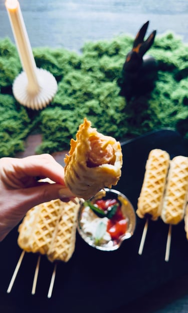 Hand holding a bitten waffle corn dog on a stick over a plate with dip and more corn dogs.