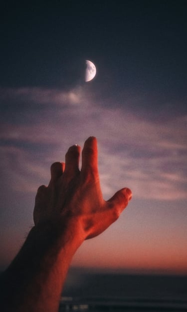 Hand reaching toward a crescent moon in a dark evening sky with sunset clouds.