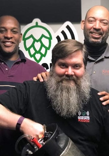 Five men posing for a photo at Beer Guys Radio with a beer keg and hop logo.