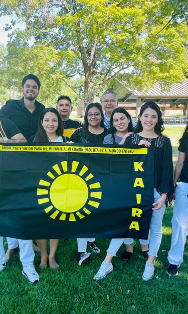 A diverse group of people posing in a park holding a black KAIRA flag with a sun logo.