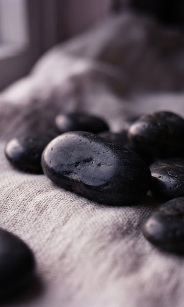 Macro shot of smooth dark basalt stones resting on textured clean linen, soft twilight window light with gentle lilac undertones, shallow depth of field.