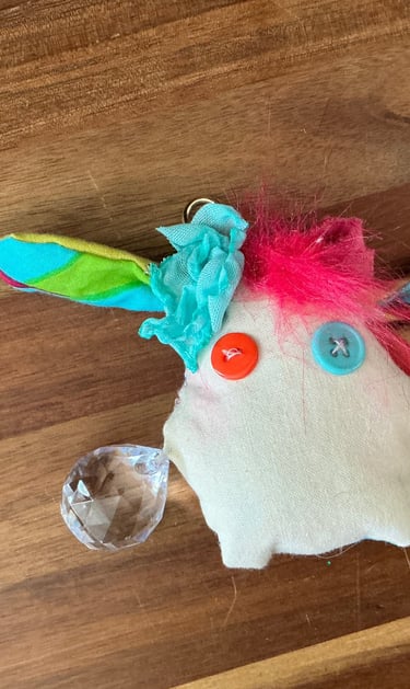 Handmade plush monster keychain with button eyes, colorful fabric ears, and a crystal charm on wood.