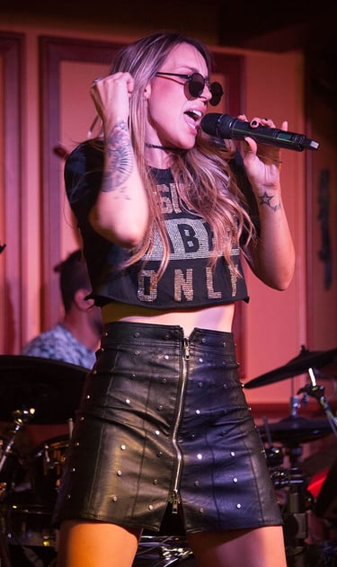 Singer Keka singing with a microphone in hand, wearing round sunglasses and a black leather skirt.