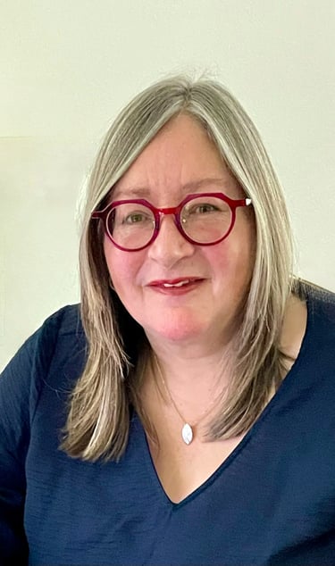 Professional portrait of a smiling woman with blonde hair wearing stylish red glasses and a blue v-neck top.