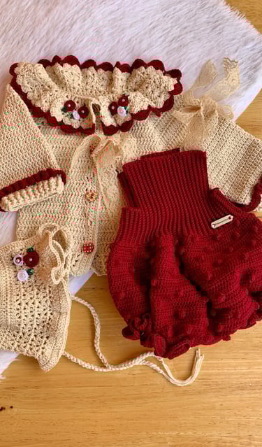 Handmade crochet baby outfit featuring a cream cardigan with floral collar, red bloomers, and a matching bonnet.