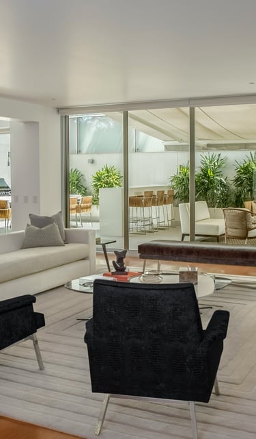 Modern luxury living room with white sofas, black velvet chairs, and sliding glass doors leading to a patio.
