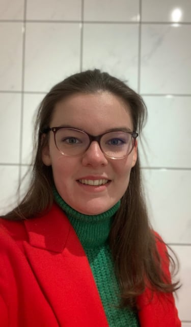 A smiling woman with glasses, wearing a red blazer and green top, standing in a bright room.