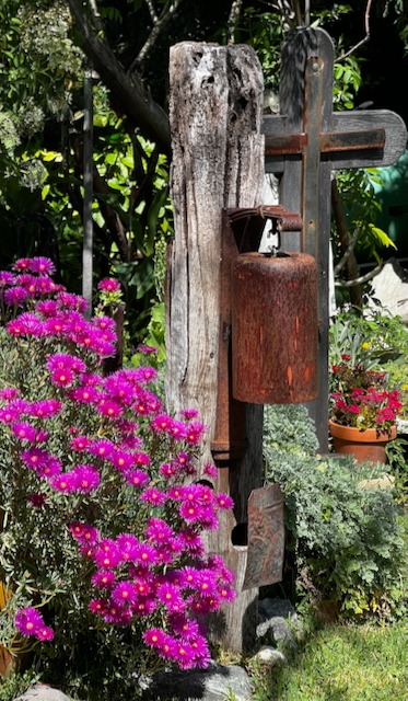Rustic metal garden bell hanging on a weathered wooden post next to bright pink ice plant flowers.