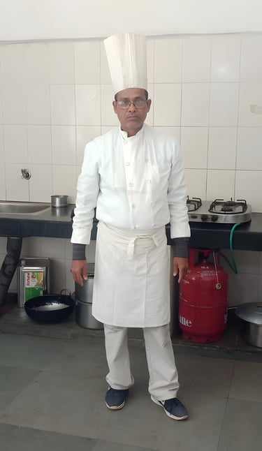 Chef in a kitchen wearing a white uniform and tall hat, standing by a countertop with a sink, stove.
