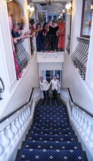 Grooms in white tuxedo jackets walking up a blue carpeted staircase at their wedding reception.