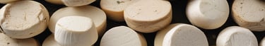 Two large, rustic wheels of cheese, one partially cut, are stacked on top of each other against a rough stone background. The cheese has a natural, textured rind and the cut reveals a smooth, pale interior.