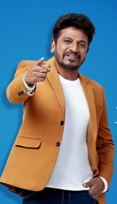 Kannada actor Shiva Rajkumar pointing forward while wearing a stylish mustard yellow blazer over a white shirt.