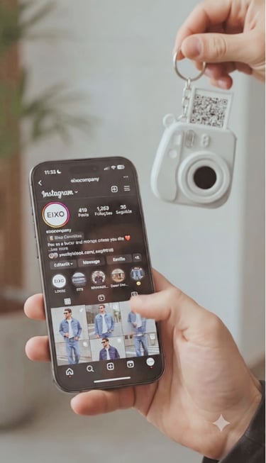 Person holding a smartphone displaying an Instagram profile next to a white polaroid camera keychain with a QR code.