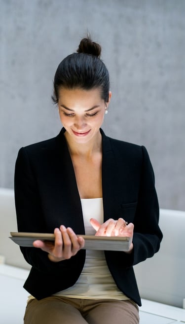 A businesswoman in a black jacket, sitting on a desk, holding a tablet computer.