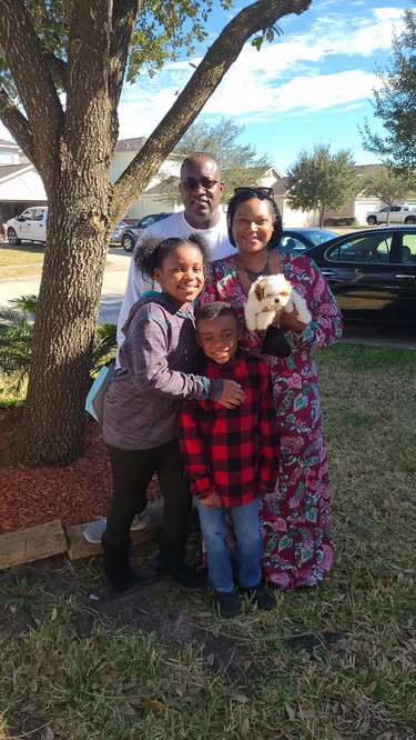 A happy family of four with their small white Mal-Shi puppy posing outdoors near a large tree.