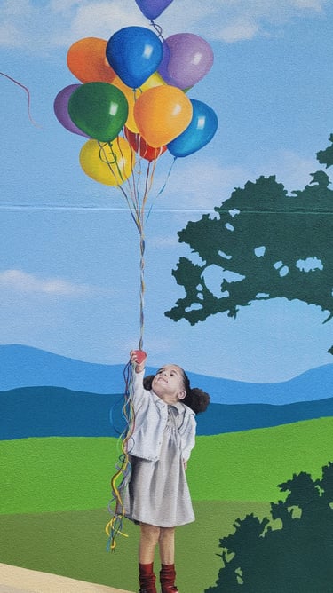 Fresque peinte sur façade. Une fillette sur un muret, tient une grappe de ballons colorés.