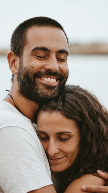 Smiling happy couple embracing outdoors with eyes closed in a romantic hug at the beach.
