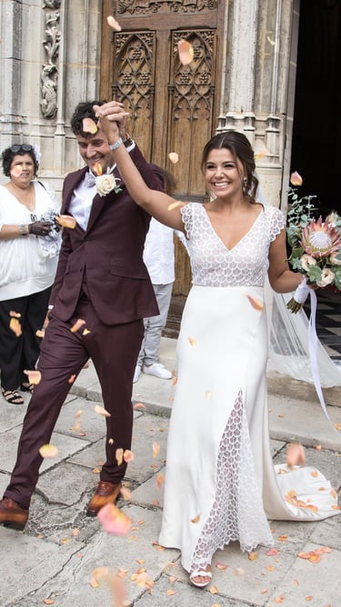 Smiling bride and groom celebrate as guests throw flower petal confetti outside a stone church.