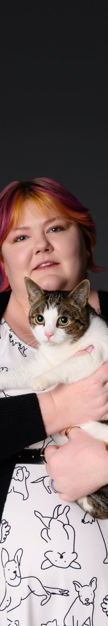 A woman with pink and orange hair holding a tabby and white cat against a dark background.