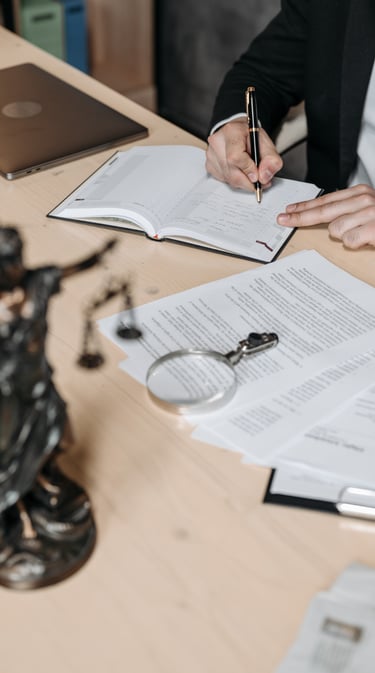 Osoba podpisująca dokument na biurku, obok otwartej książki, leży lupa oraz figura Temidy, w tle lap