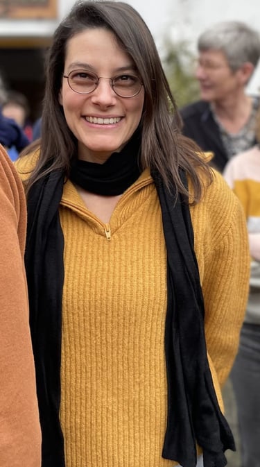 Julia Braun lächelt mit Brille, ockerfarbenem Wollpullover und schwarzem Schal