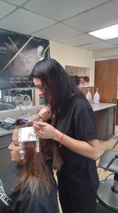 A student at a beauty school applying hair highlights with foil to a client in a salon setting.