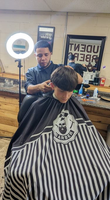 A student barber giving a haircut with electric clippers in a barber college setting.