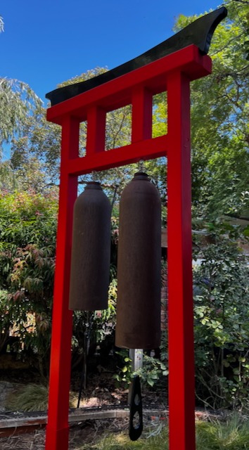 Red Japanese torii gate wind chime featuring two large brown bells in a lush green garden.