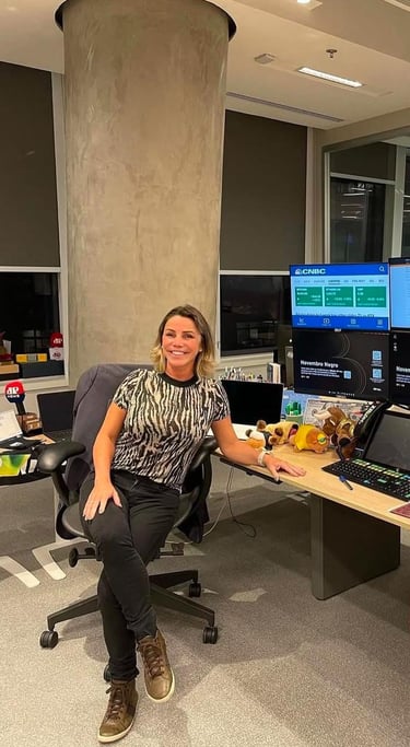 A smiling woman in a tiger-print top and black jeans sits at a trading desk with multiple CNBC screens, a Novenbro Negro