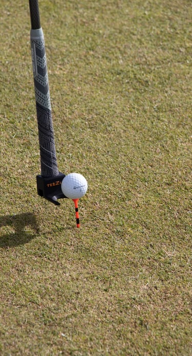 A golf club with a 'TeeZUP' attachment holding an orange tee and a golf ball, set up on a grassy course, ready for a shot