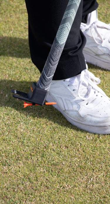 Person picking up a golf tee of the grass using the TeeZUP tool on grass, with a club and white sneakers visible.
