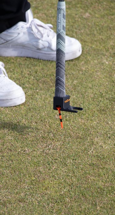 A golfer uses the 'TeeZUP' tool, attached to the end of a golf club, to pick up a orange tee in place on the grass