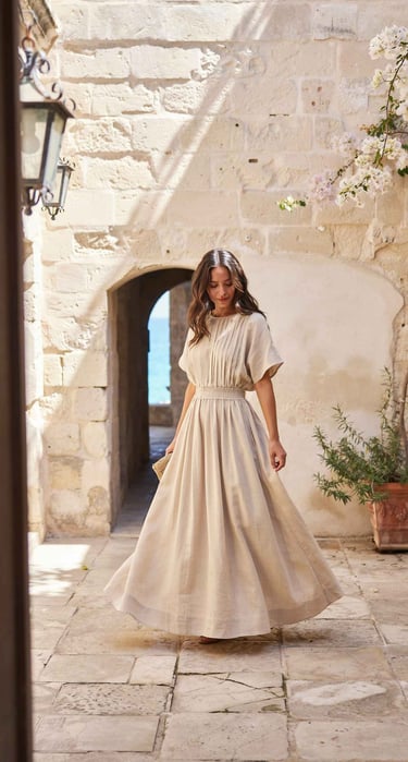 Woman in a beige linen maxi dress walking through a sunlit Mediterranean stone courtyard.
