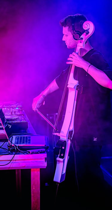 A male musician performs a live electronic set playing an electric cello with DJ equipment under purple stage lights.