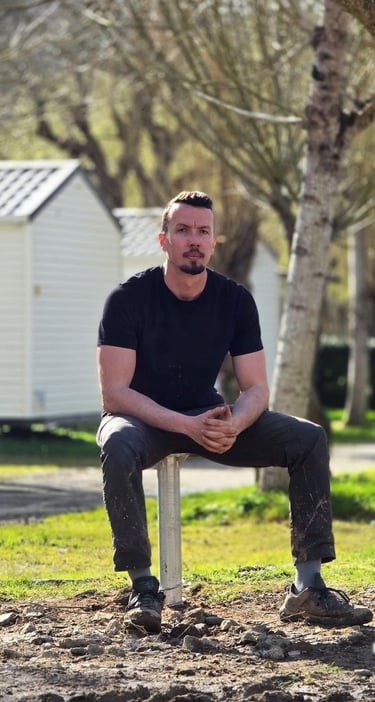 Man in black t-shirt sitting outdoors at a campground with mobile homes and trees in the background.
