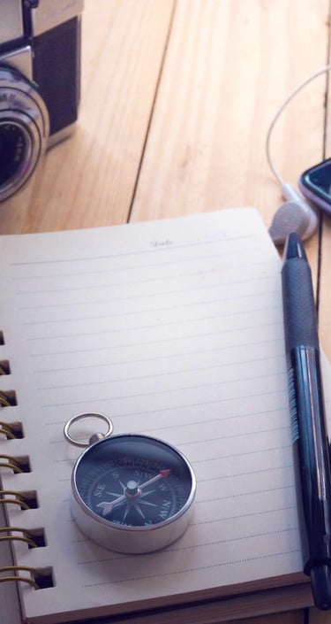 A compass on top of a notebook, with a pen and camera on the side