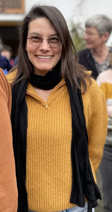 Julia Braun lächelt mit Brille, ockerfarbenem Wollpullover und schwarzem Schal
