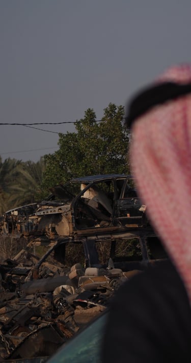 an arab man in a red and white scarf looking at a destroyed car in the by the palm trees in Iraq