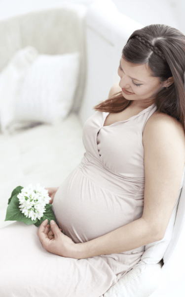 A smiling pregnant woman sitting on a white chair holding white flowers while wearing a beige dress.