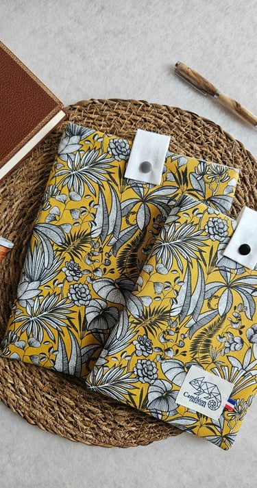 Yellow floral fabric book sleeves with a vintage journal and wooden pen on a woven mat.