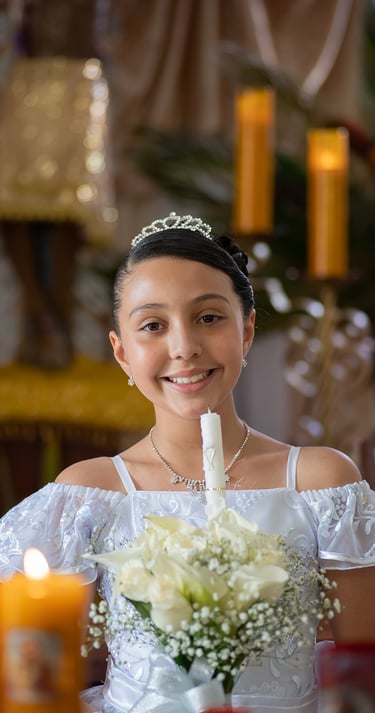 Niña sonriente con vestido blanco de Primera Comunión y tiara sosteniendo una vela ceremonial y un r