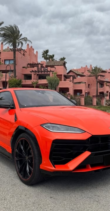 Bright orange Lamborghini Urus luxury SUV parked in front of a pink Mediterranean villa with palm trees.