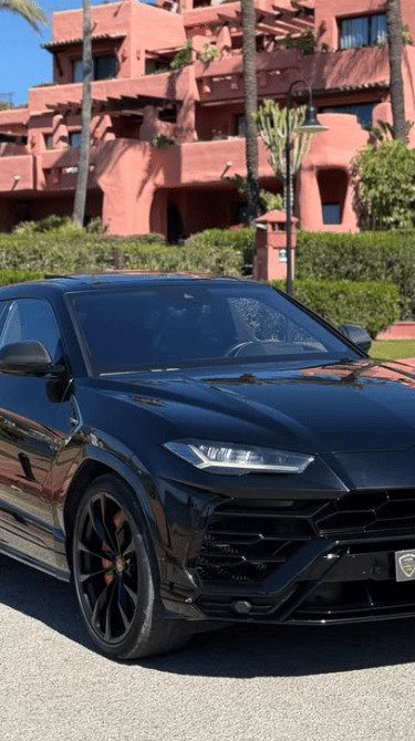 Black Lamborghini Urus luxury SUV for rent parked in front of a Mediterranean resort with palm trees.