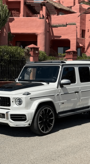 White Brabus Mercedes G-Wagon luxury SUV parked in front of pink Mediterranean villas for rent.