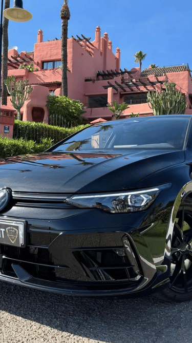 Black Volkswagen Golf R rental car parked in front of a luxury pink resort with palm trees.