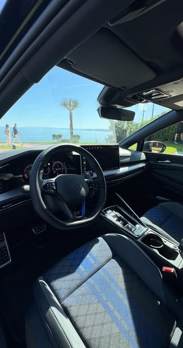 Interior view of a modern Volkswagen Golf R cockpit with digital display and blue patterned seats parked near a beach.