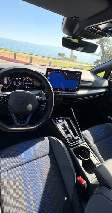 Interior view of a Volkswagen Golf R featuring a digital cockpit and blue plaid seats parked by the ocean.