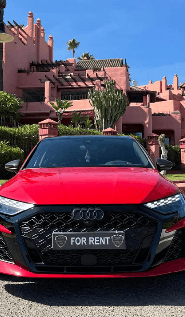 A red Audi luxury car rental parked in front of a Mediterranean style resort on a sunny day.
